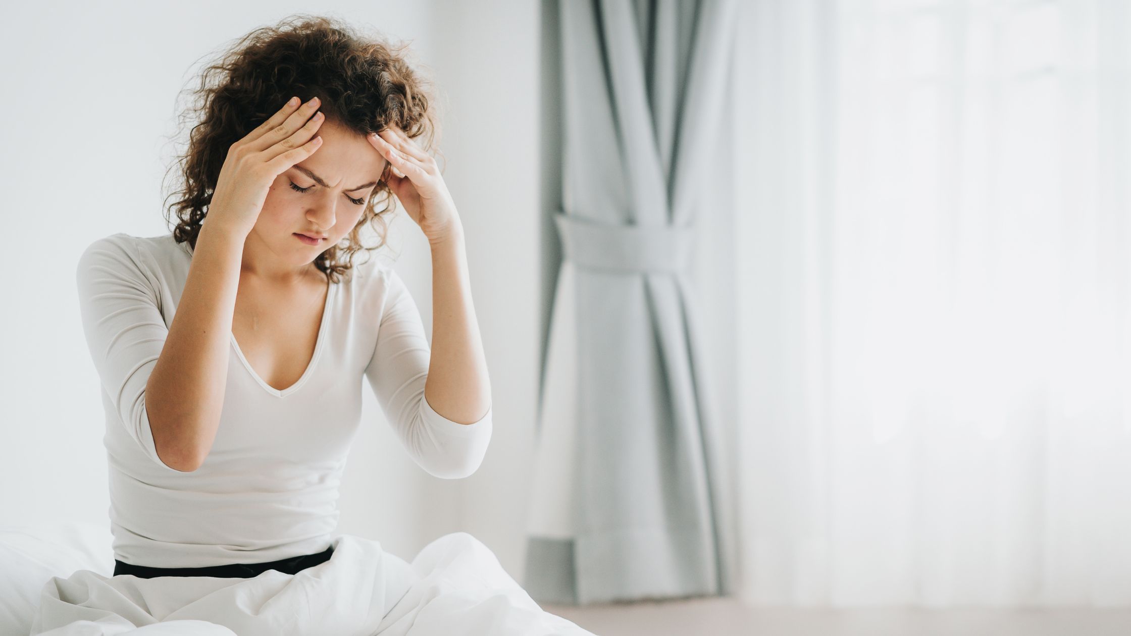 Woman sitting in bed holding her head in discomfort, possibly from a headache.