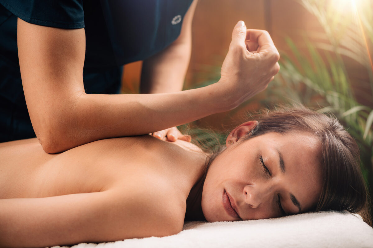 Close-up of a woman lying face down on a massage table with eyes closed as a massage therapist applies firm elbow pressure to release tension in her upper back.