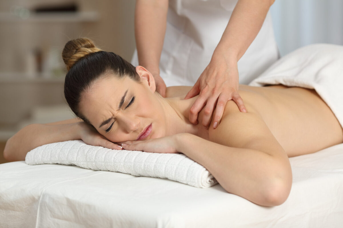 Massage therapist applying firm pressure to a woman’s upper back and shoulders during a deep tissue massage, woman appears to feel intense sensation.