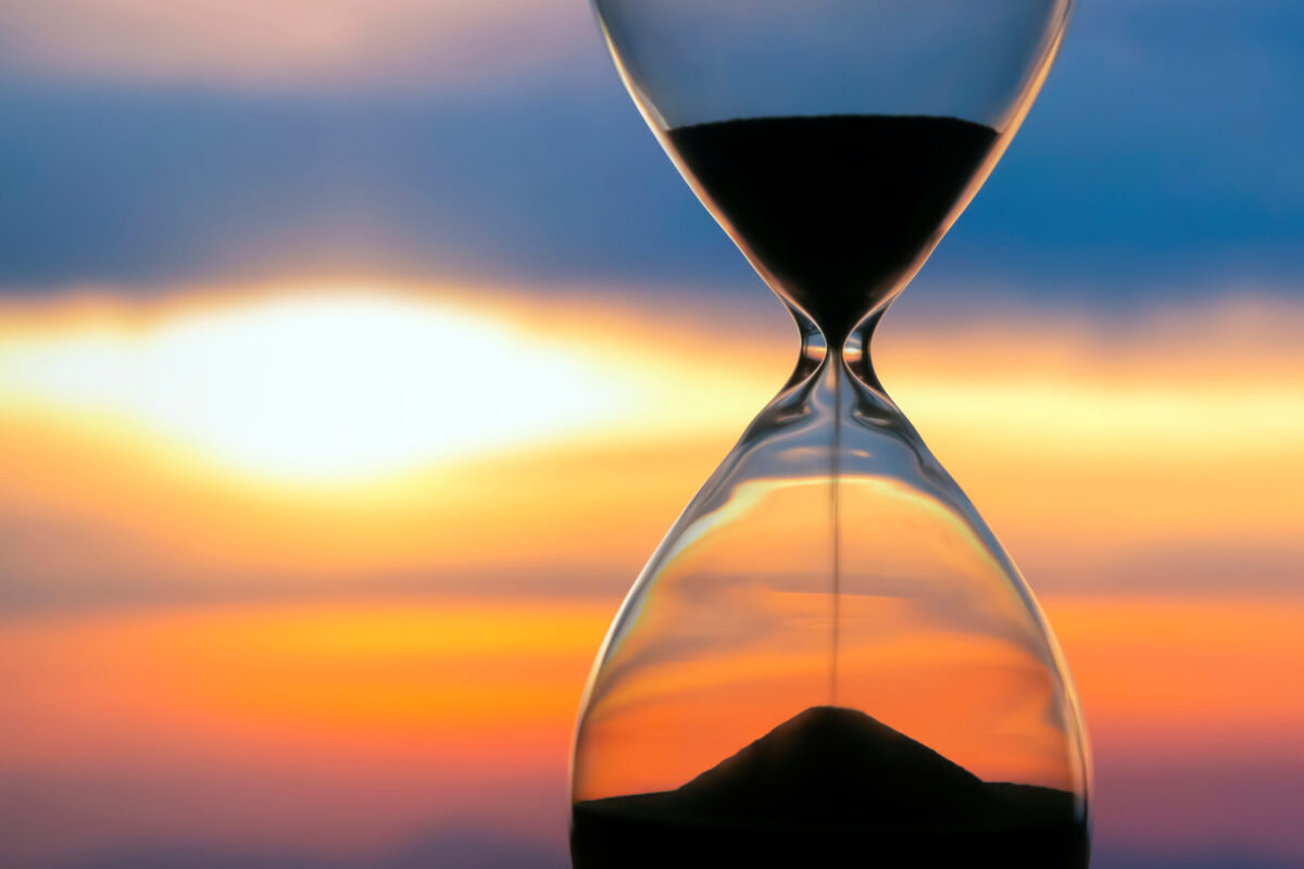 Close-up of an hourglass with black sand running through it, silhouetted against a vivid orange, yellow, and blue sunset sky.