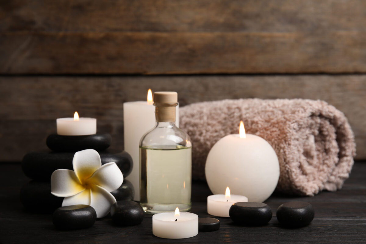 Lit candles, massage oil, black stones, white flower, and rolled towel in a spa setting.