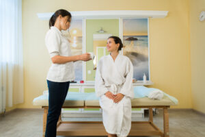 Woman in a white robe relaxing in a spa massage room as an attendant hands her a glass of water