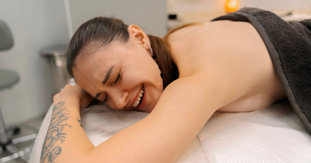A woman lies face down on a massage table, partially covered with a dark towel, grimacing and reacting to the strong pressure of an intense deep tissue massage.