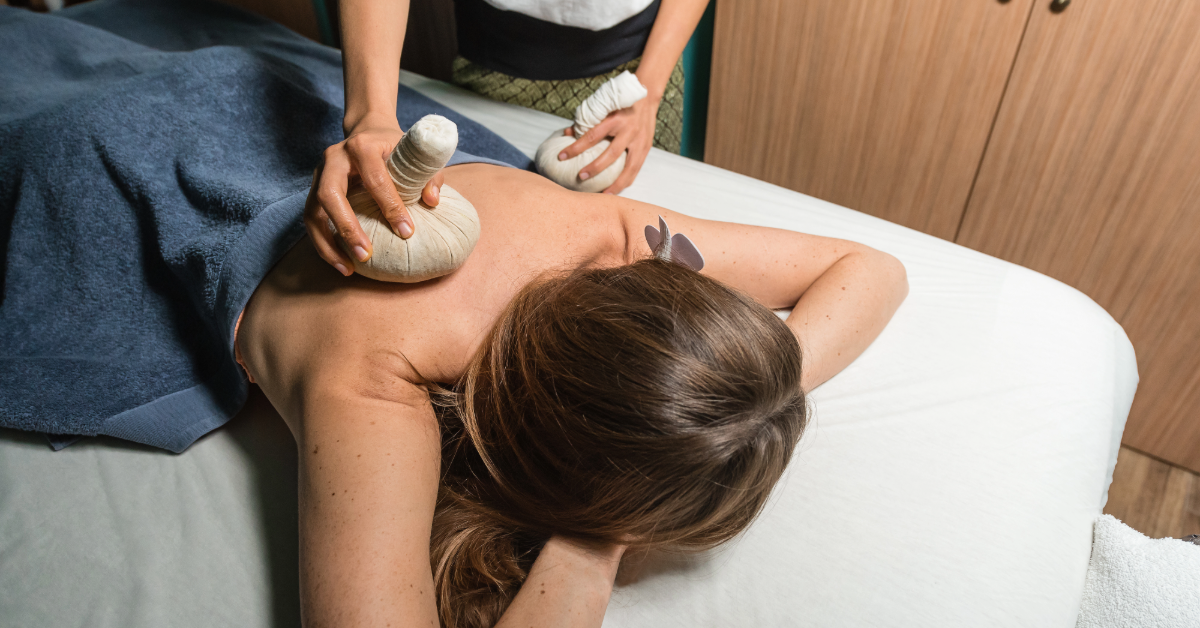 A massage therapist applies warm herbal compresses to a woman’s shoulder and back as she lies face down on a spa table, using traditional techniques for muscle relaxation and stress relief.
