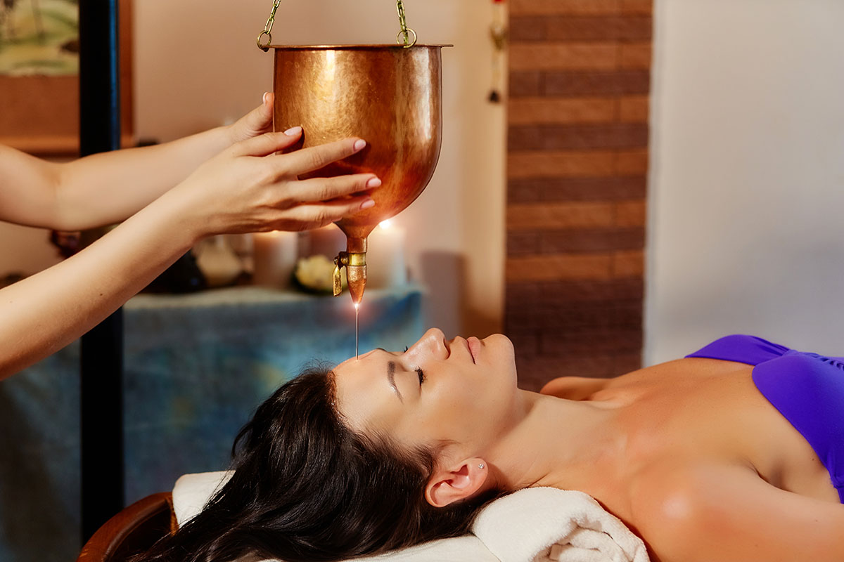 Therapist performing Shirodhara, gently pouring a continuous stream of warm oil onto a client’s forehead during an Ayurvedic treatment.