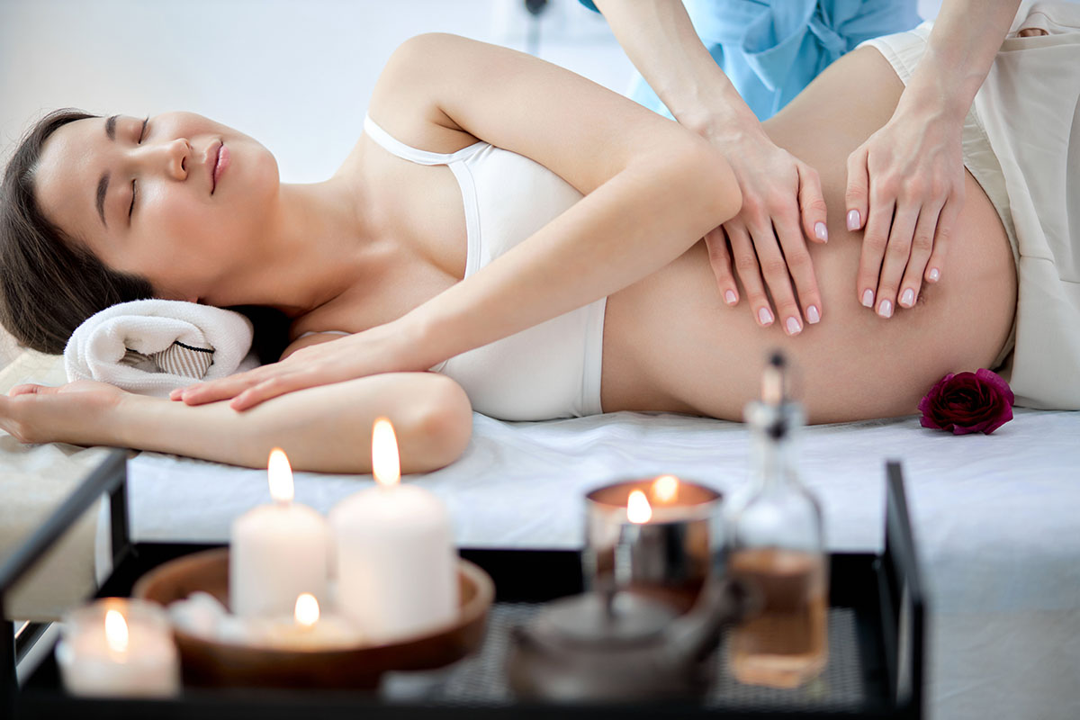 Pregnant woman lying on her side receiving a gentle belly massage in a serene spa setting with lit candles and massage oils nearby.