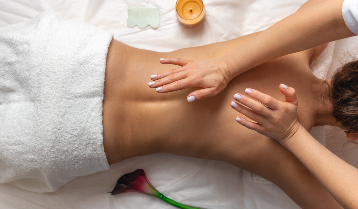 Massage therapist using both hands to perform a back massage on a client lying under a white towel, with a candle and flower nearby.