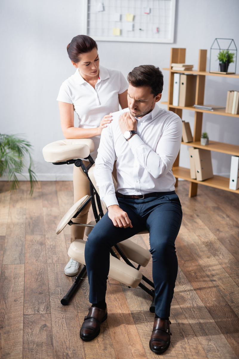 Female therapist consulting a male office worker about shoulder pain beside a massage chair