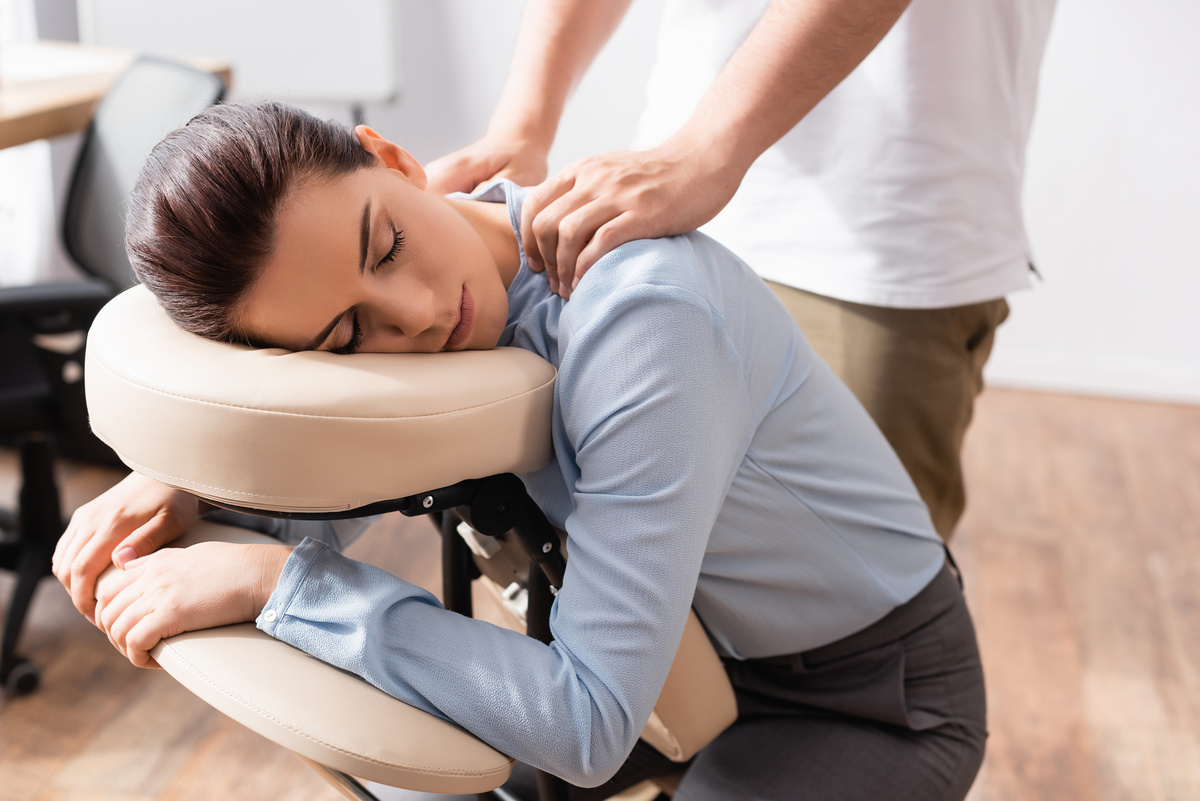 Female employee resting on a massage chair face cradle while therapist applies shoulder pressure in an office