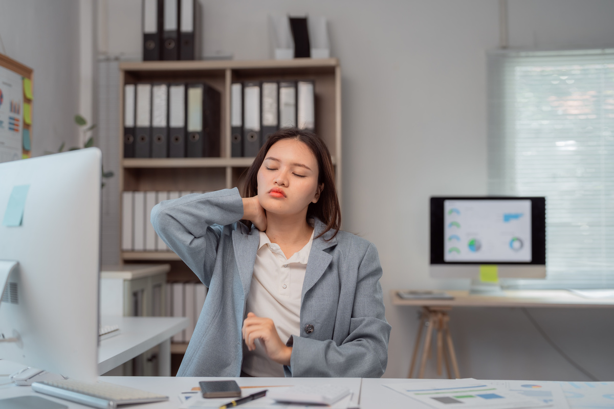 Female office worker in blazer holding her neck in pain while working at desk